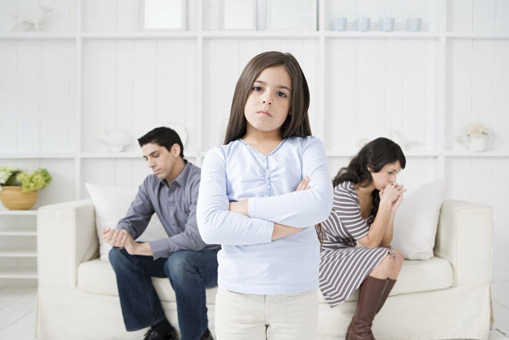Young girl stands arms crossed with parents in the background looking distressed