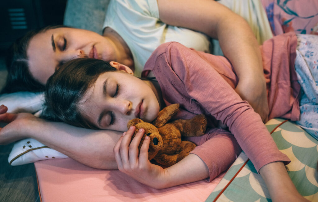 Portrait of serene daughter embracing a teddy bear lying next to mom over mats at night. Close up of woman sleeping and hugging little girl with closed eyes. Security and family protection concept.