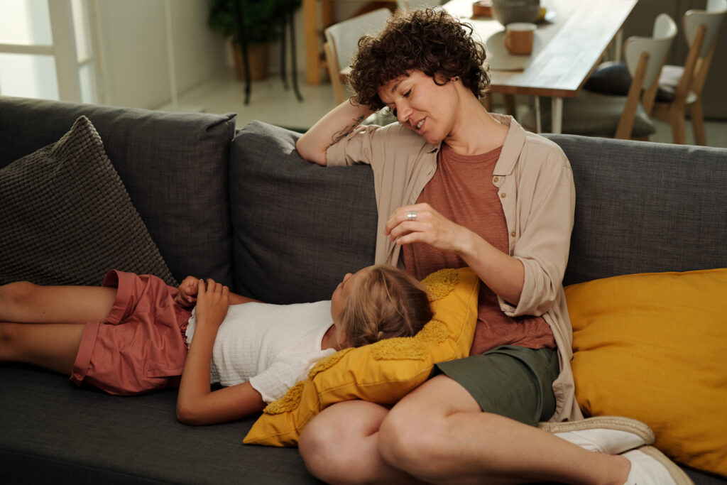 Youthful serene girl lying on pillow on her mother knees and both having chat while relaxing on comfortable couch in living room